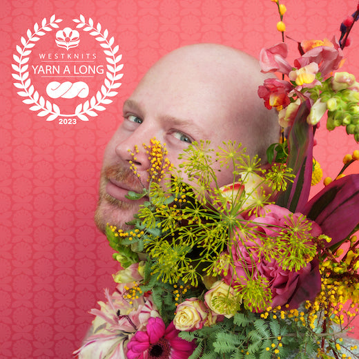 Photo of Stephen holding a bouquet of pink and purple flowers. He is staring in the camera with a playful smile. He is against a pink background decorated with flowers. A white Westknits YAL logo is on the top left corner.