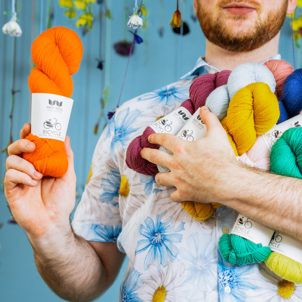 Stephen holds an armfull of bright skeins of West Wool. He has a skein of Beatrix, our power orange in one hand and is standing against a pale blue background.