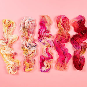 Five wavy skeins of Hedgehog Fibre yarn in red, pink, and yellow on a pink background