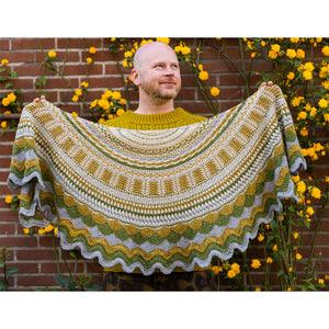 This image shows Stephen West holding a sample of the Achievement Badge Shawl, part of the Westknits 2025 YAL. The sample is beautifully knitted in West wool yarn in blue, ochre, green, and cream shades. He stands on a flowery background.