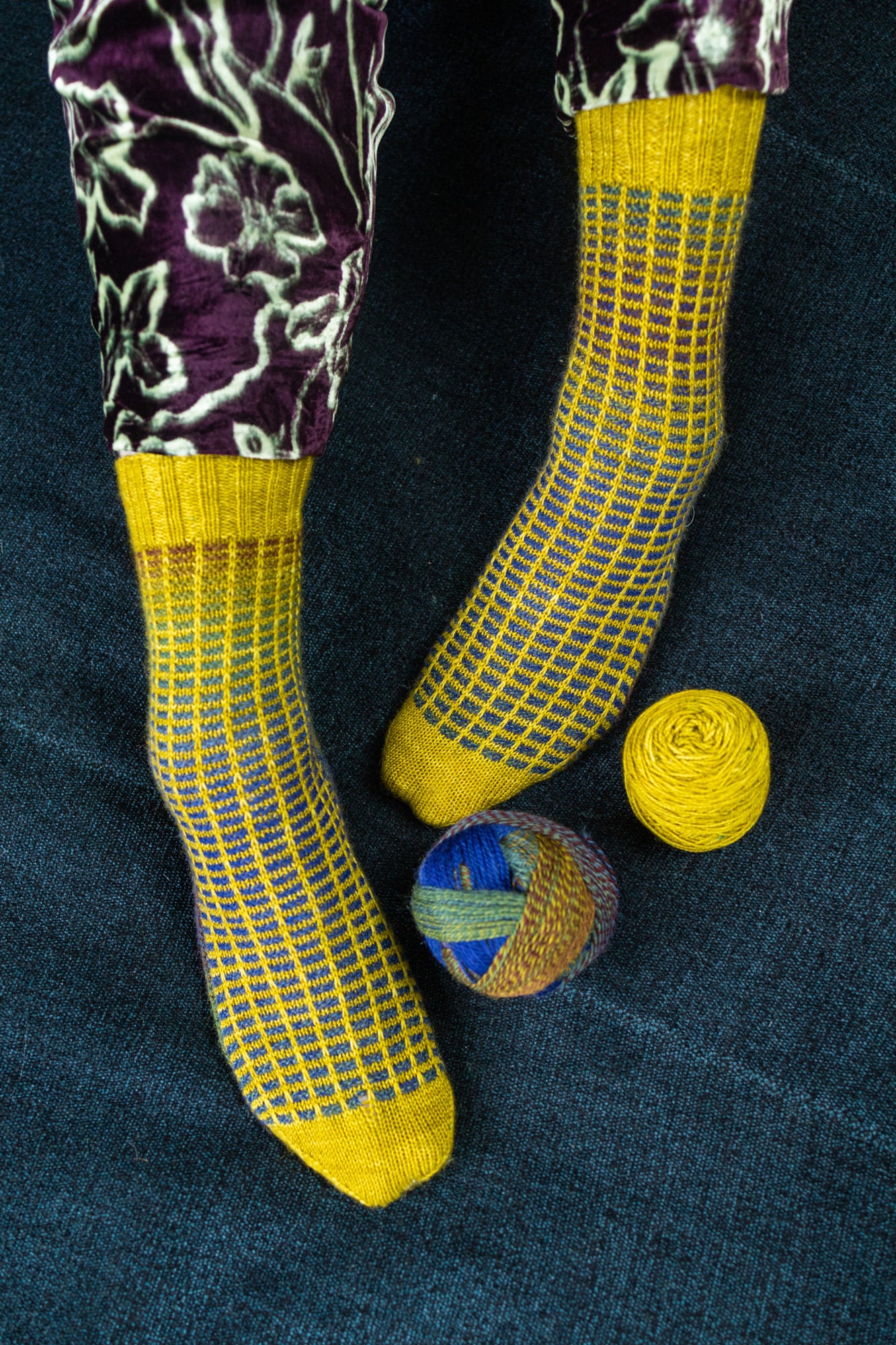 This image shows Stephen West wearing a yellow and blue sample of the Botanic Socks by Westknits. He stands on a denim blue background next to two balls of yarn.