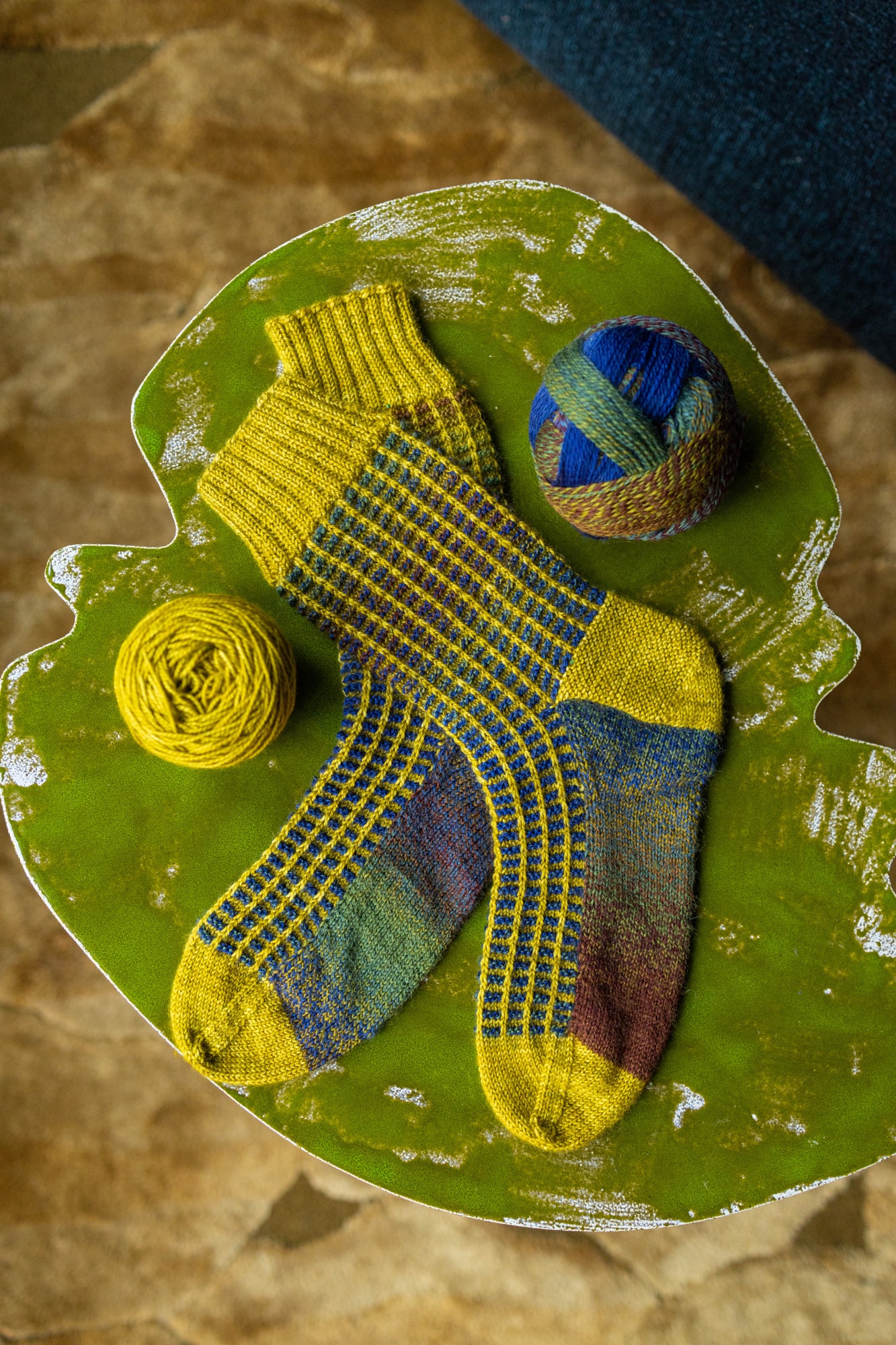 This image shows a yellow and blue sample of the Botanic Socks by Westknits. He stands on a green small table next to two balls of yarn.