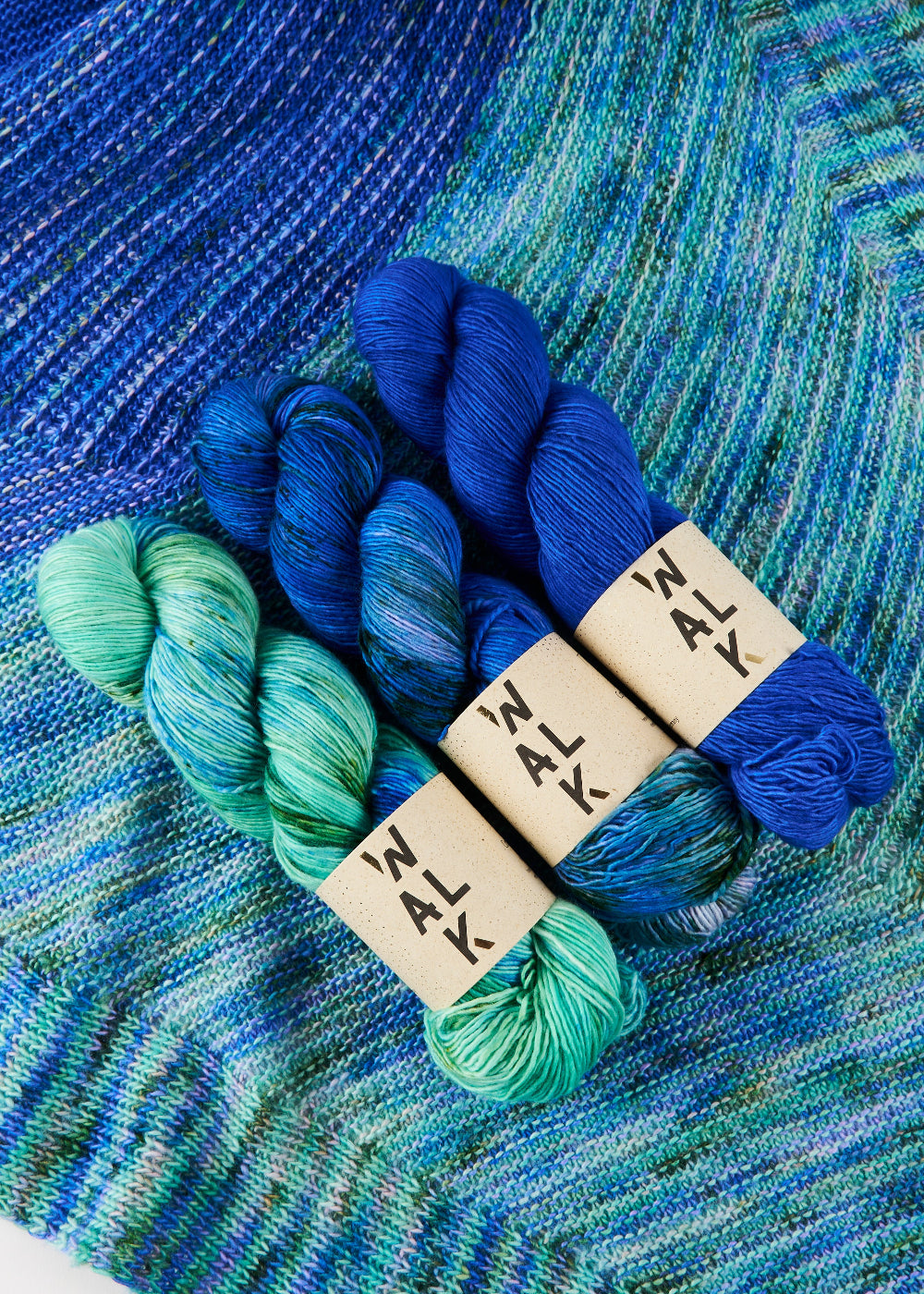 Three skeins of blue and green yarn with 'Walk' labels on a knitted blue and green shawl background.