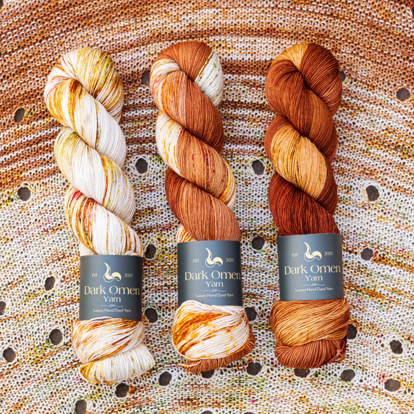 This image shows three skeins of Dark Omen Yarn in brown shades, on a beautifully knitted sample of the Camp Craft Shawl.