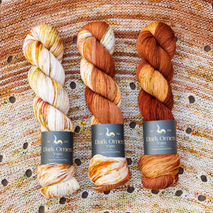 This image shows three skeins of Dark Omen Yarn in brown shades, on a beautifully knitted sample of the Camp Craft Shawl.