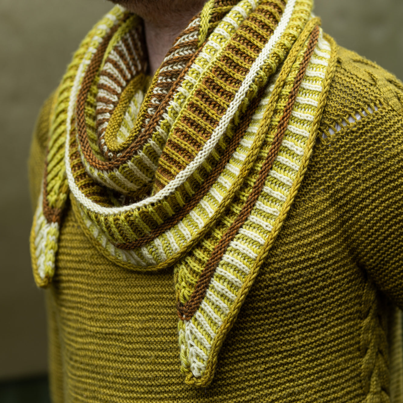 This image shows a close-up of Stephen West wearing a sample of the Cool Column Collected Shawl by Westknits. The sample is beautifully knitted in green, brown, and yellow tones.