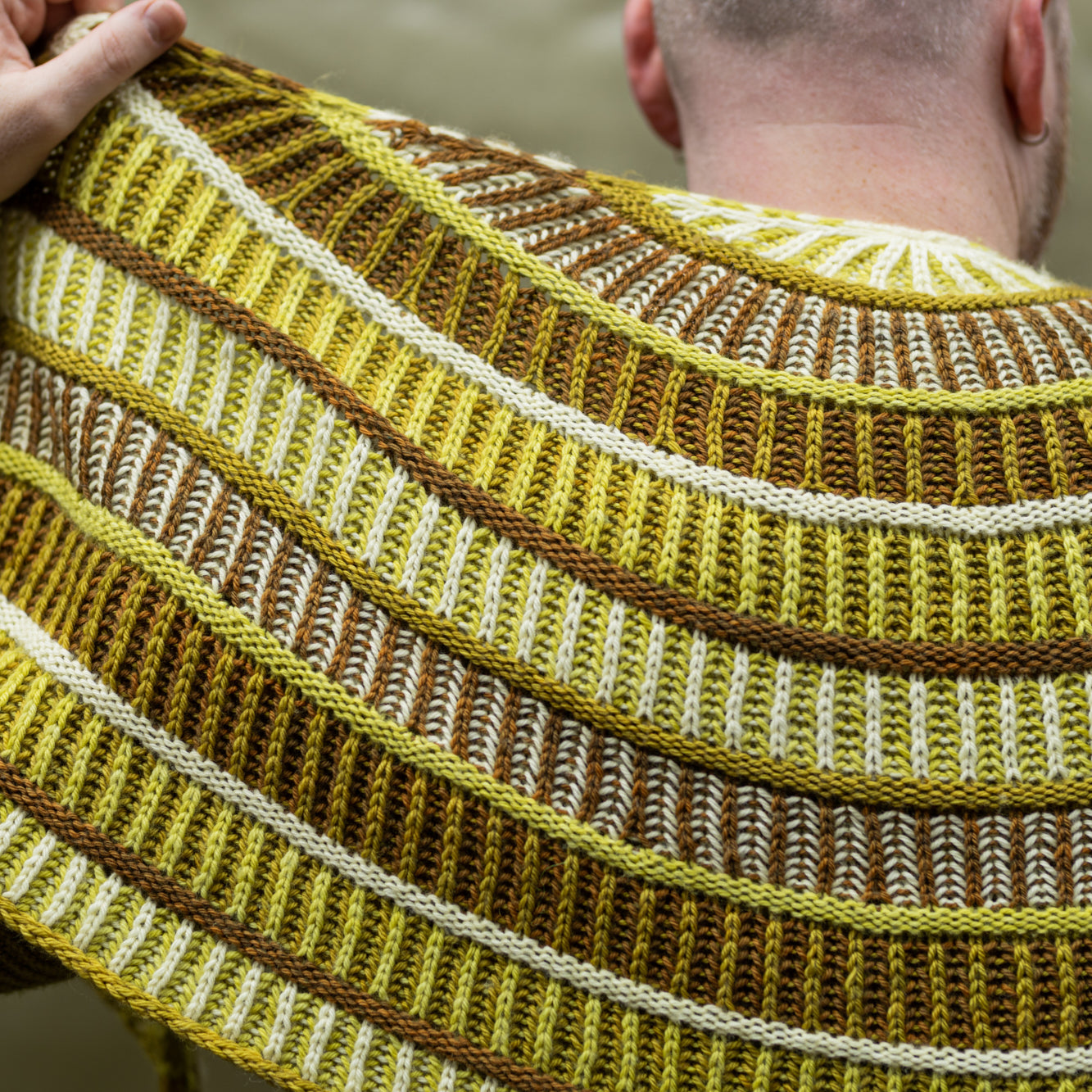 This image shows a close-up of Stephen West wearing a sample of the Cool Column Collected Shawl by Westknits. The sample is beautifully knitted in green, brown, and yellow tones.