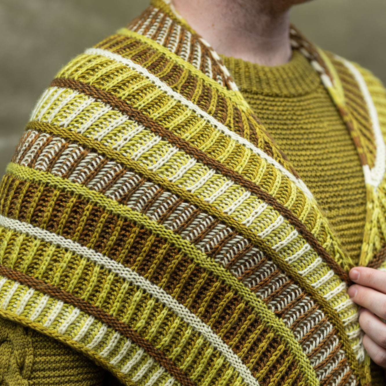 This image shows a close-up of Stephen West wearing a sample of the Cool Column Collected Shawl by Westknits. The sample is beautifully knitted in green, brown, and yellow tones.