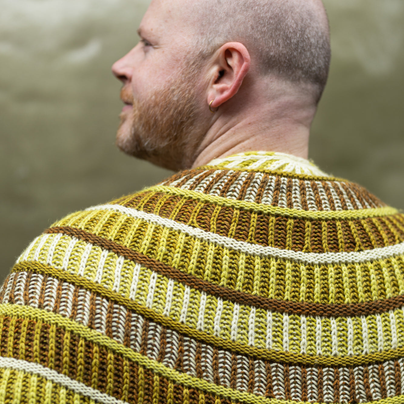 This image shows Stephen West holding a sample of the Cool Column Collected Shawl by Westknits. The sample is beautifully knitted in green, brown, and yellow tones.