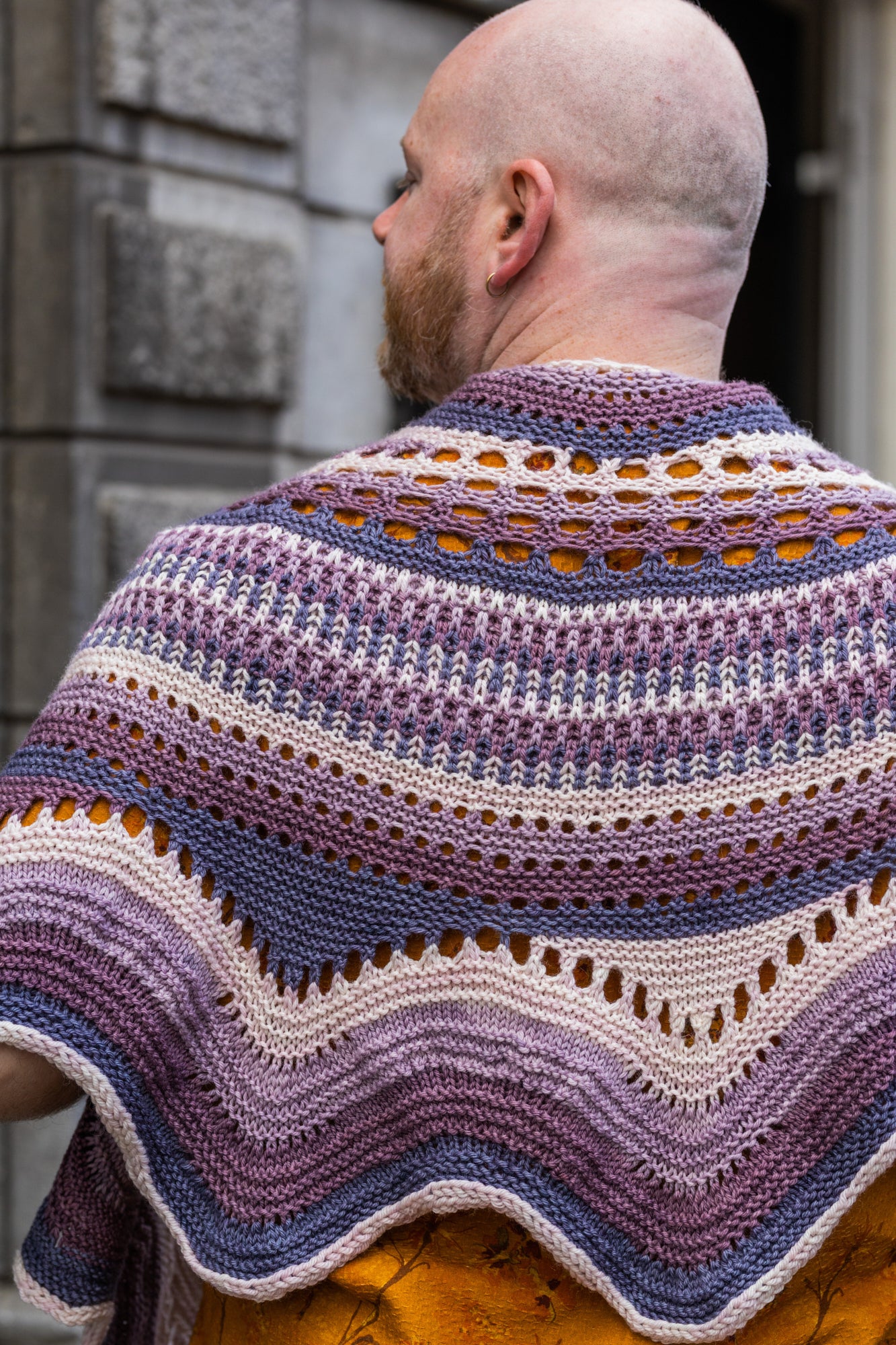 This image shows Stephen West holding a sample of the Eyelet Journey Shawl by Westknits. The sample is knitted in purple and pink gradients. The background shows a building in Amsterdam.