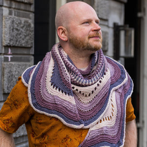 This image shows Stephen West holding a sample of the Eyelet Journey Shawl by Westknits. The sample is knitted in purple and pink gradients. The background shows a building in Amsterdam.