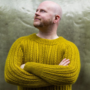 This image shows Stephen West waring a beautifully knitted sample of the Fir Rib Sweater on a green background.
