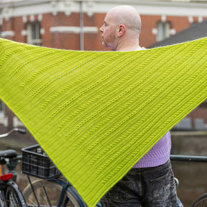 This image shows Stephen West wearing a beautifully knitted shawl. The shawl is the Fir Rib Shawl and it is knitted in a bright green colour. The background shows a building in Amsterdam.