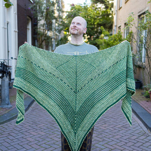 This image shows Stephen West holding the Húsafell Shawl. The shawl is knitted with green tones. The background shows a street in Amsterdam centre.