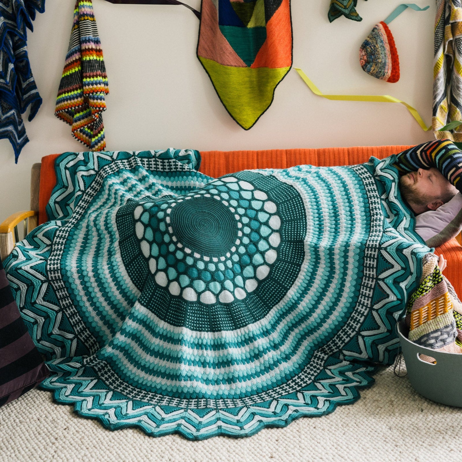 This image shows Stephen West sleeping under a beautifully knitted sample of the Slipstravaganza Blanket in cool blue and turquoise tones.