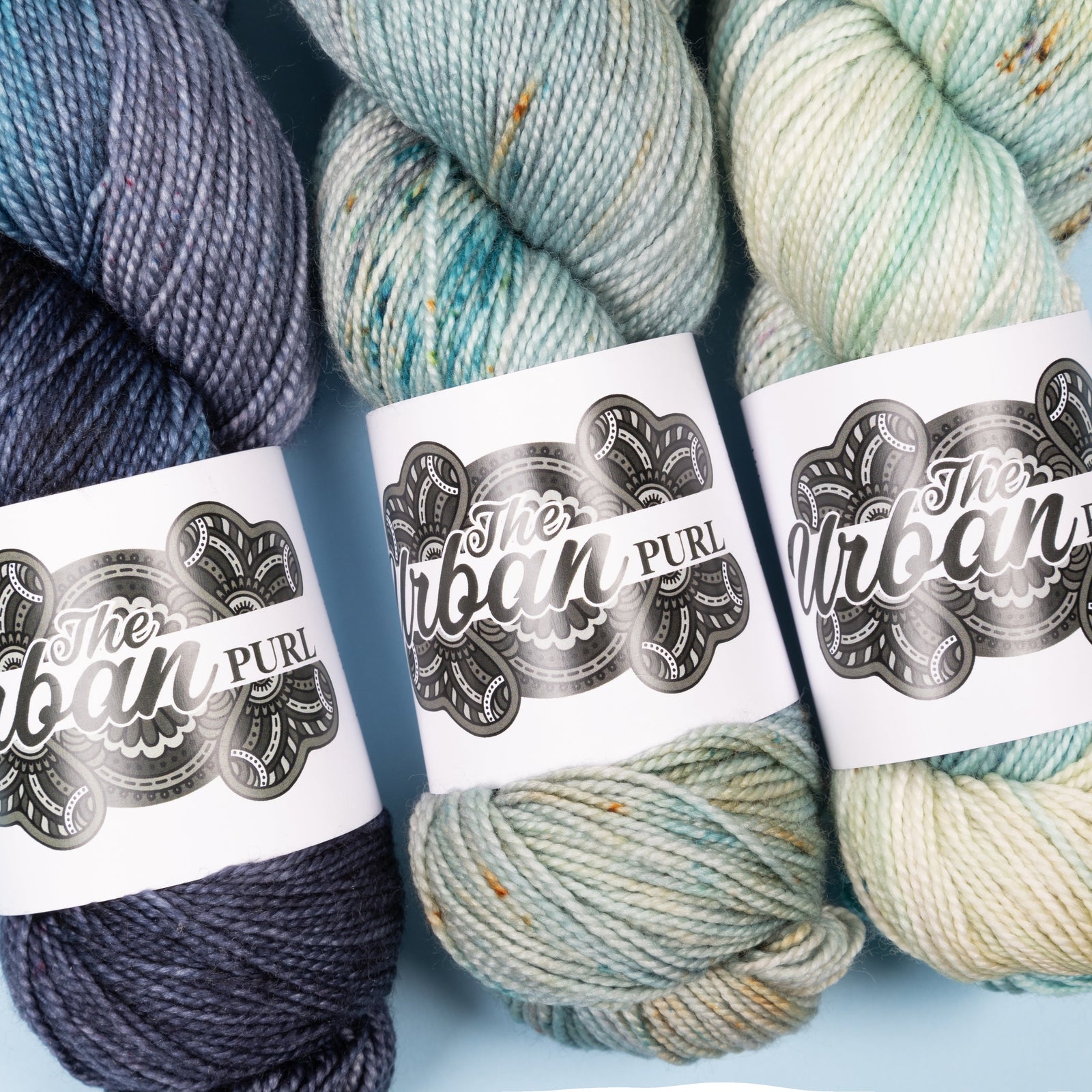This image shows a close-up of 3 skeins of Lux High Twist from the Urban Purl in blue tones, on a light blue background.