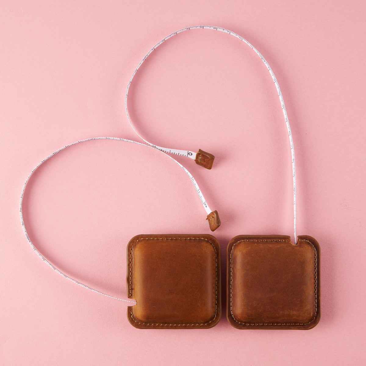 Two brown leather measuring tapes with a white tape on a pink background