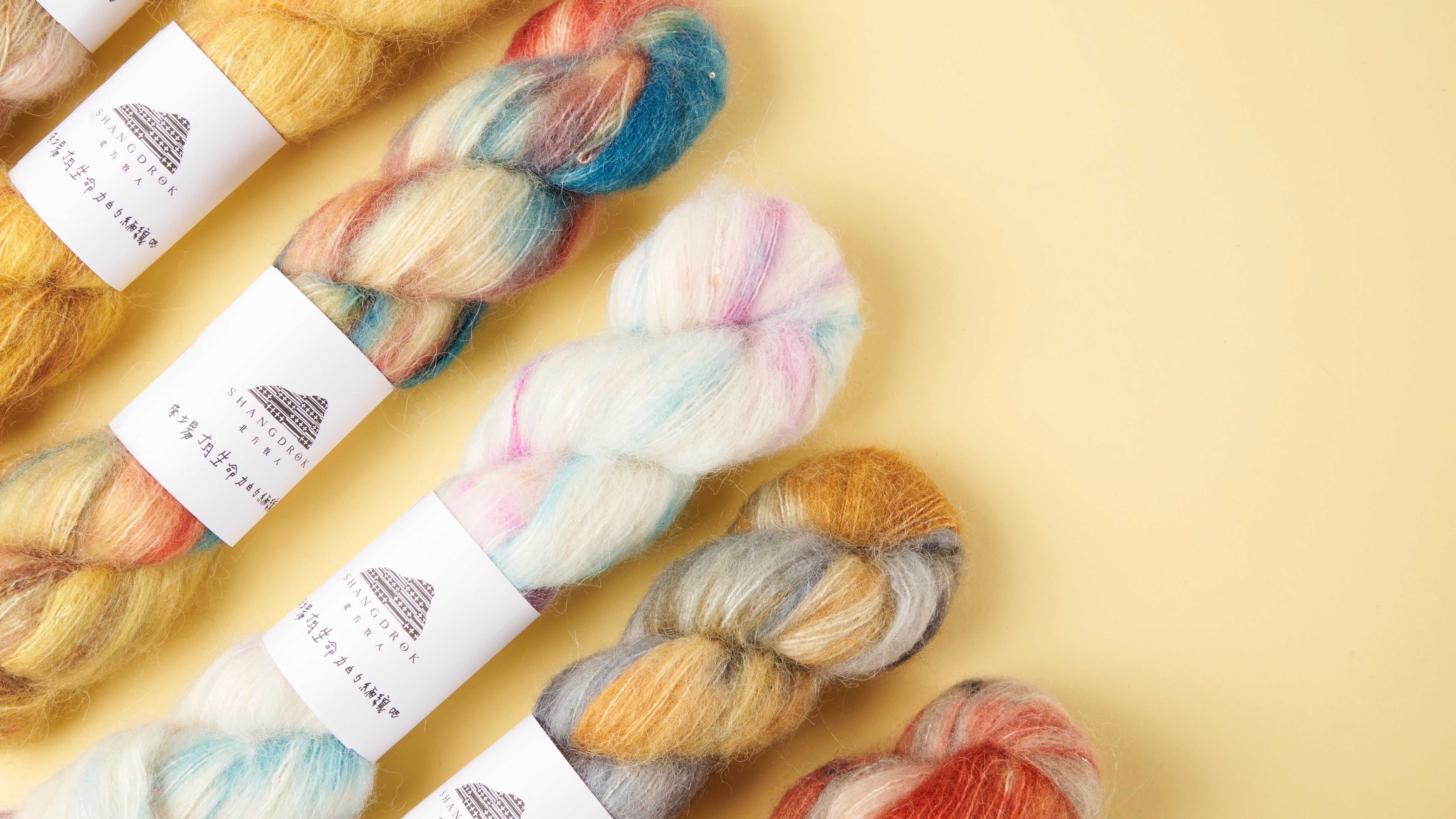 Five skeins of alpaca shimmer lace from Shangdrok ln a pale yellow background. The yarns display a mix of colors including yellow, blue, pink, orange, gray, and white, each with a white paper label.