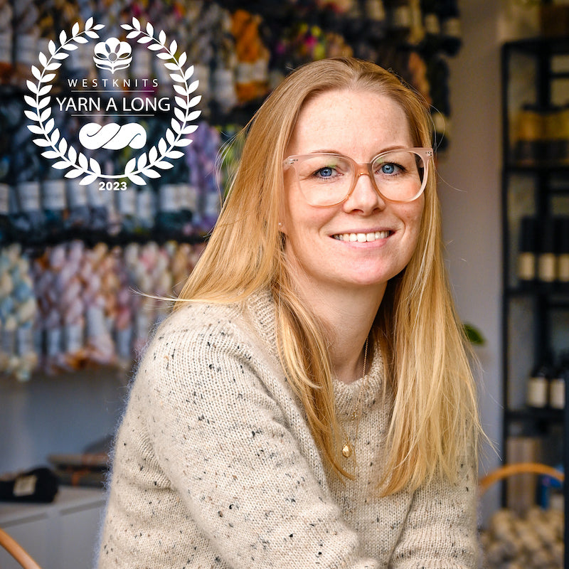 Image shows Malene sat and watching in the camera. She is in front of a wall full of Sysleriget yarn. She is wearing a beige speckled knit sweater. A white Westknits YAL logo is on the top left corner.