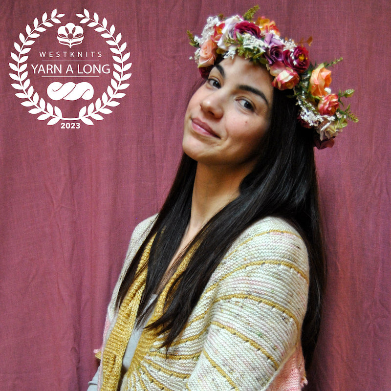 Photo of Natasha against a light burgundy background. She has an orange, red, and purple flower crown on her head and is wearing a Doodler Shawl by Stephen West. A white Westknits YAL logo is on the top left corner.