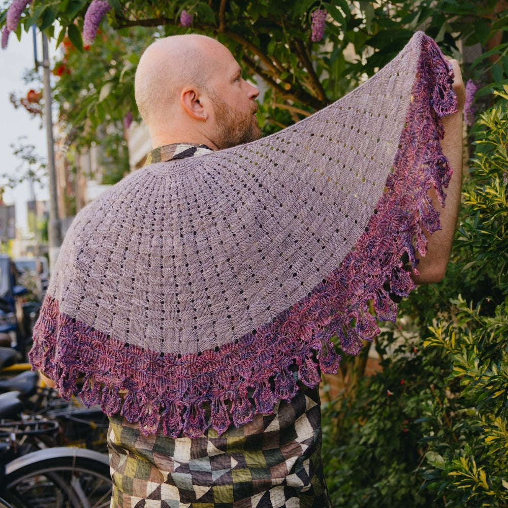 Stephen West outdoors wearing a large knitted shawl draped over his shoulders, with bicycles and greenery in the background.