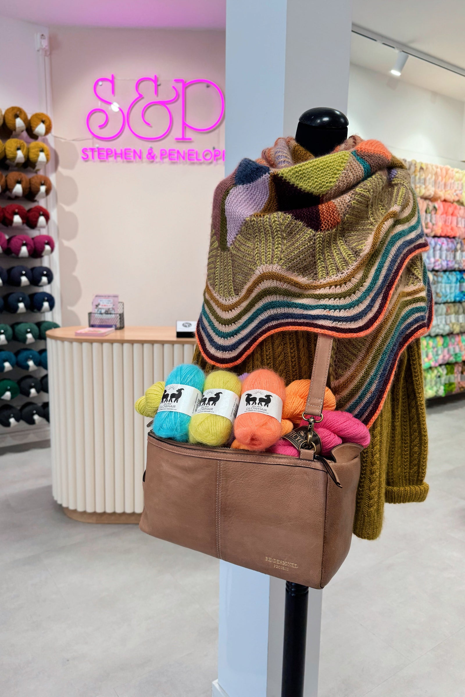 Colorful knitted shawl draped over a mannequin with yarn balls and a brown bag in a store setting.