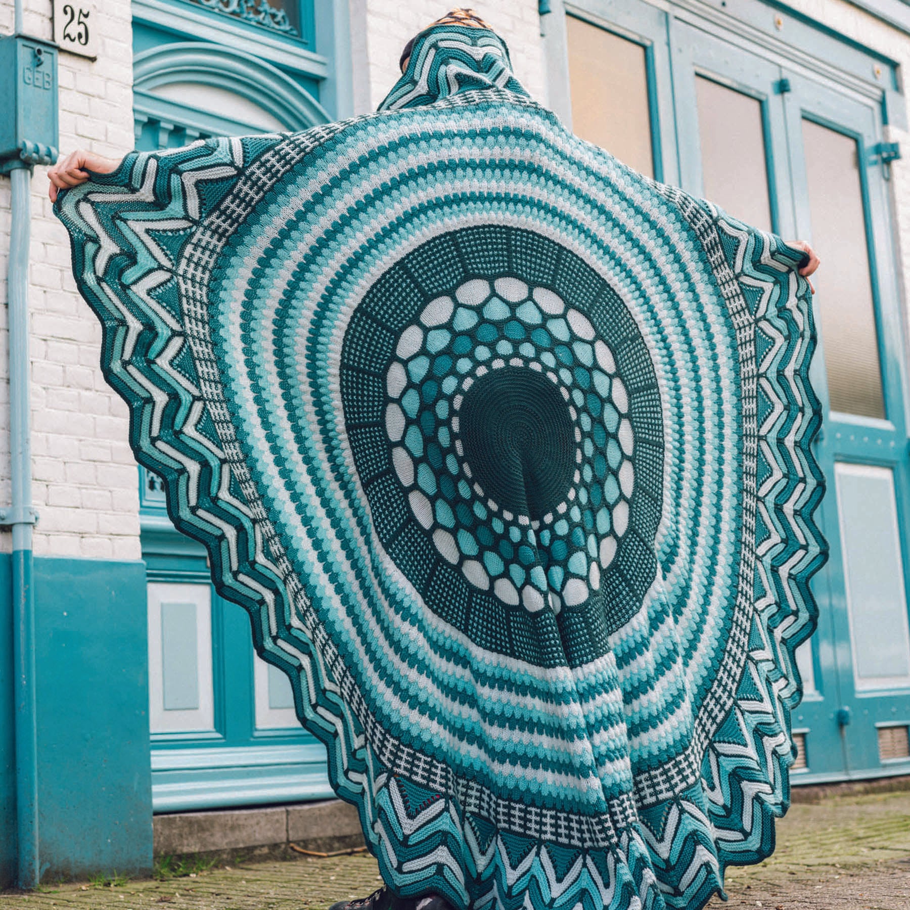 This image shows Stephen West wearing a beautifully knitted sample of the Slipstravaganza Blanket in cool blue and turquoise tones. 