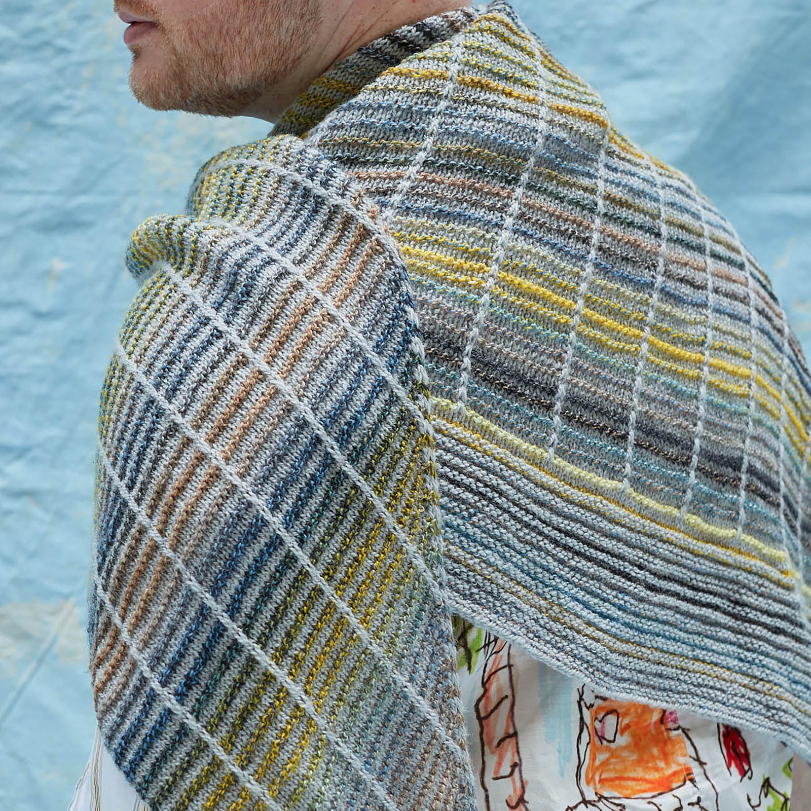 This image shows Stephen West wearing a beautifully knitted sample of the Botanic Shawl by Westknits around his neck. The sample is knitted in yellow, grey, and blue pastel colours. It stands on a light blue background. 