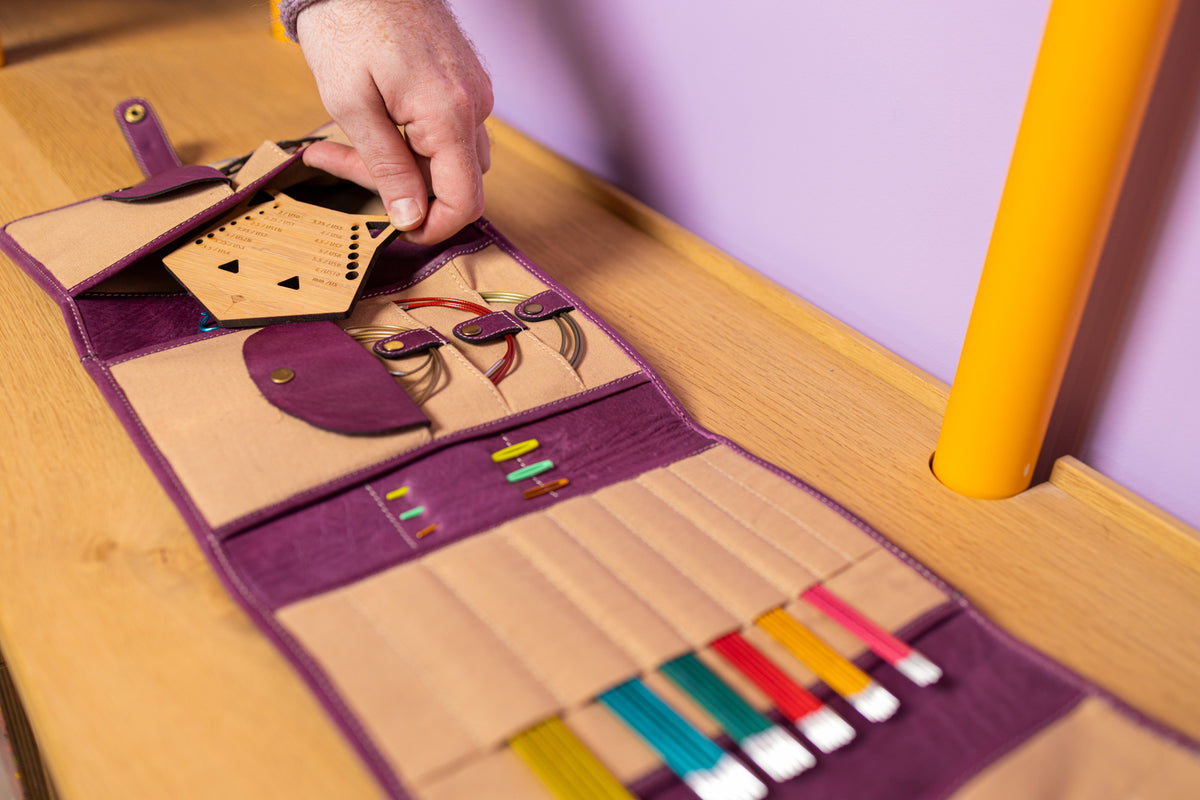This image shows Project 14 - Aubergine, filled with needles and notions. The bag is part of a collaboration between Re:Designed and Westknits. Stephen West is holding a needle gauge out of the bag.