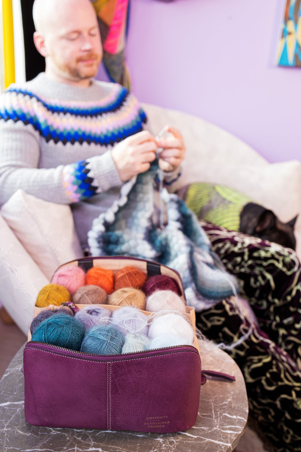 This image shows Project 9 - Aubergine, filled with yarn. The bag is part of a collaboration between Re:Designed and Westknits. Stephen West is on the background knitting on a sofa. 