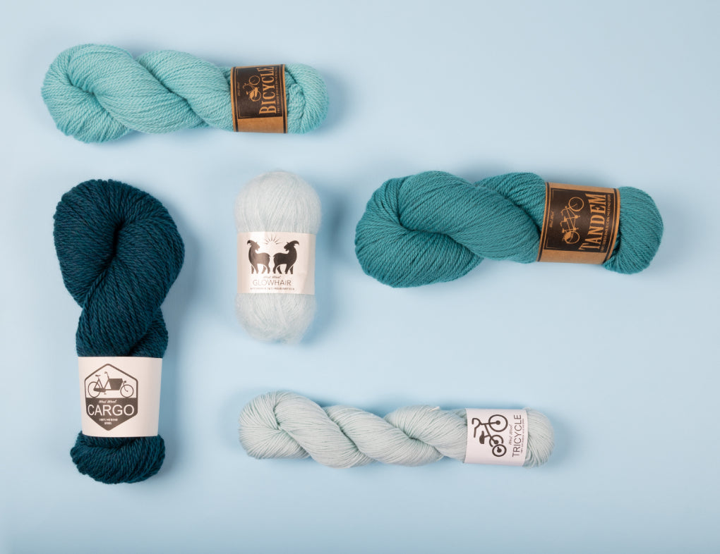 This image shows 5 skeins of West Wool yarn in blue gradient on a light blue background. Each skein is in a different shade of blue and of a different base (Tandem, Cargo, Tricycle, Bicycle, Glowhair). 