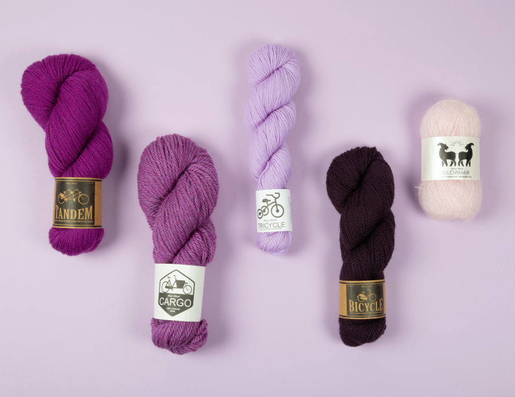 This image shows 5 skeins of West Wool yarn in purple gradient on a lilac background. Each skein is in a different shade of purple and of a different base (Tandem, Cargo, Tricycle, Bicycle, Glowhair). 