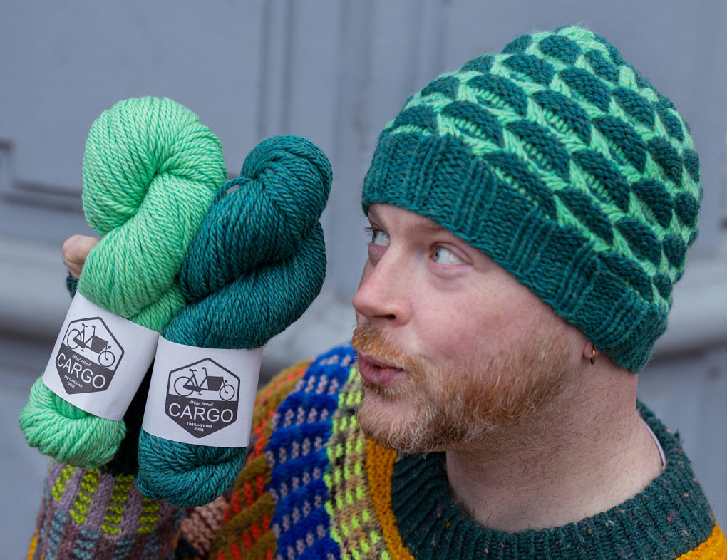 The image shows Stephen West wearing a beautifully knitted sample of the Stony Stream Hat in two contrasting greens, while holding two green skeins of West Wool chunky weight Cargo. The background shows a fading building detail. 
