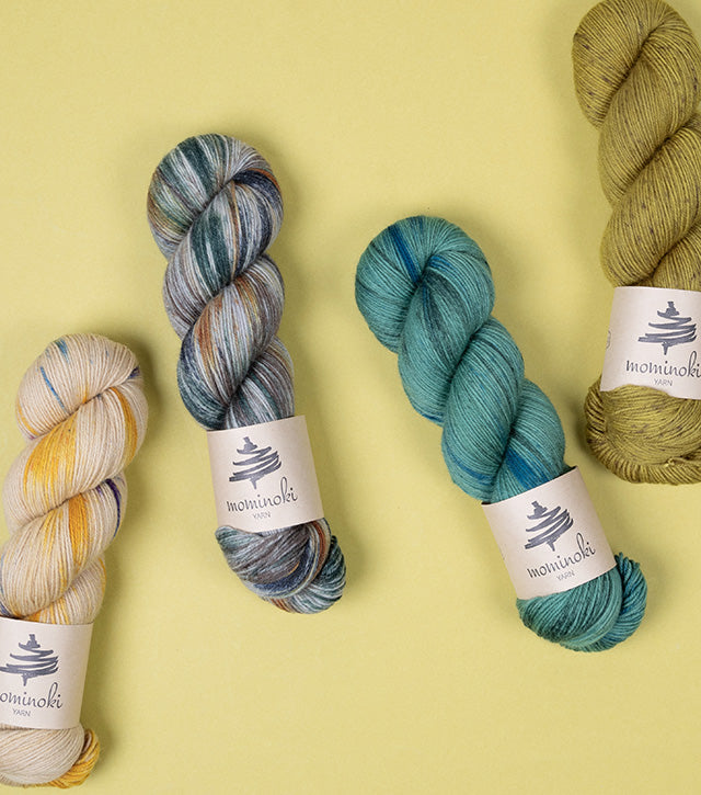 This image shows 5 skeins of Mominoki Yarn in various shades of green on a light green background. 