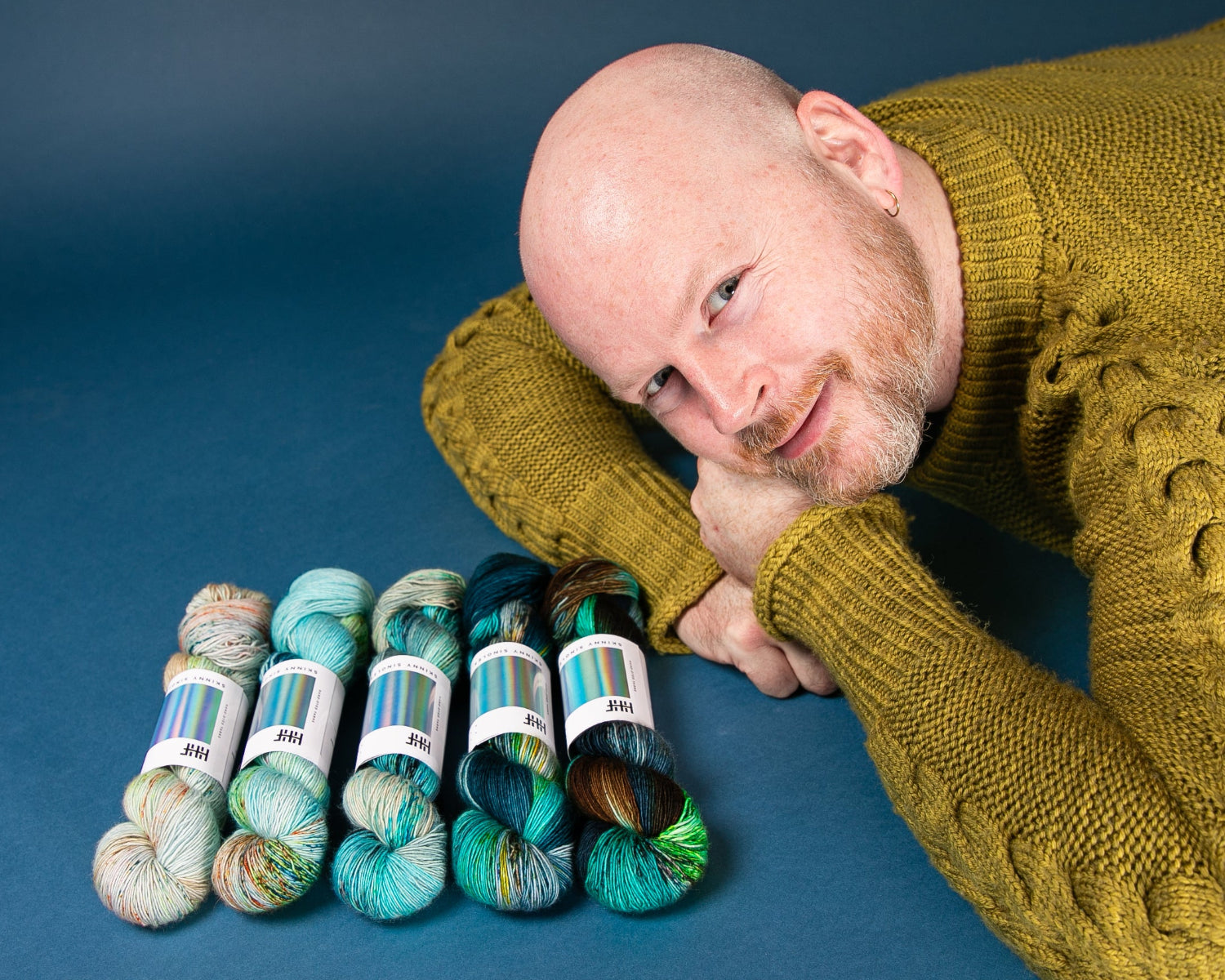 Stephen West  in a mustard yellow sweater with skeins of yarn on a blue background