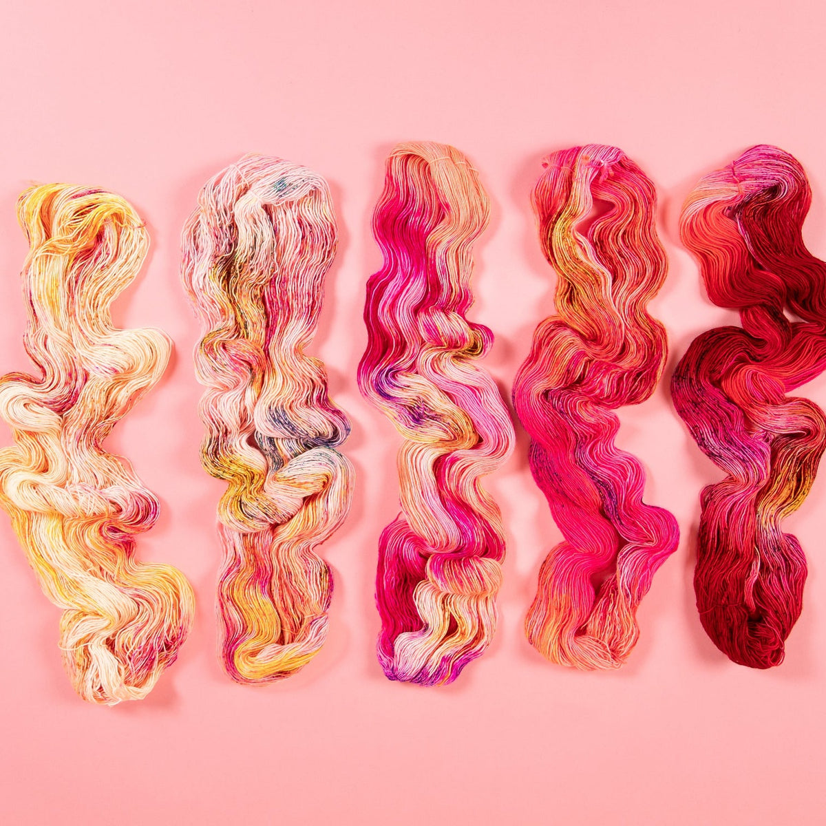 Five wavy skeins of Hedgehog Fibre yarn in red, pink, and yellow on a pink background