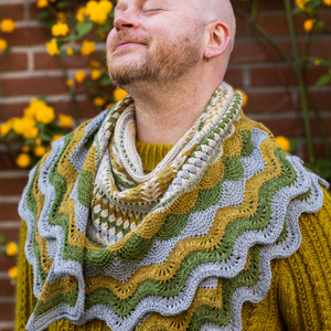 Stephen West wearing a knitted sample of the YAL 2025 shawl in blue, yellow, green and cream tones