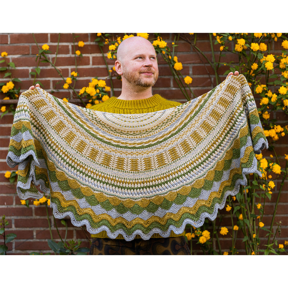 This image shows Stephen West holding a sample of the Achievement Badge Shawl, part of the Westknits 2025 YAL. The sample is beautifully knitted in West wool yarn in blue, ochre, green, and cream shades. He stands on a flowery background. 