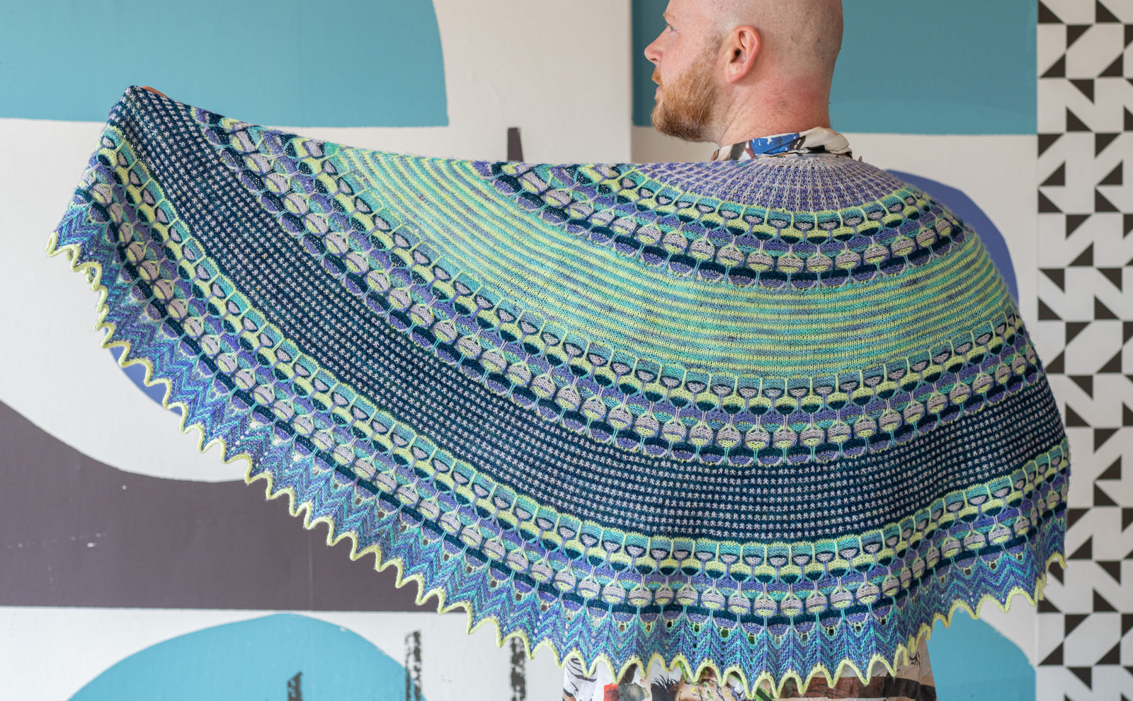 This image shows Stephen West wearing a beautifully knitted sample of the Aurora Cabin Shawl. The sample is knitted in blue, turquoise and purple shades. He stands on a colourful background. 