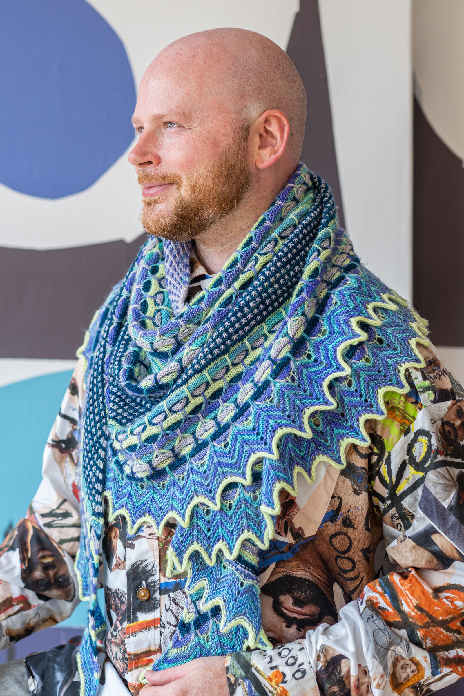 This image shows Stephen West wearing a beautifully knitted sample of the Aurora Cabin Shawl. The sample is knitted in blue, turquoise and purple shades. He stands on a colourful background. 