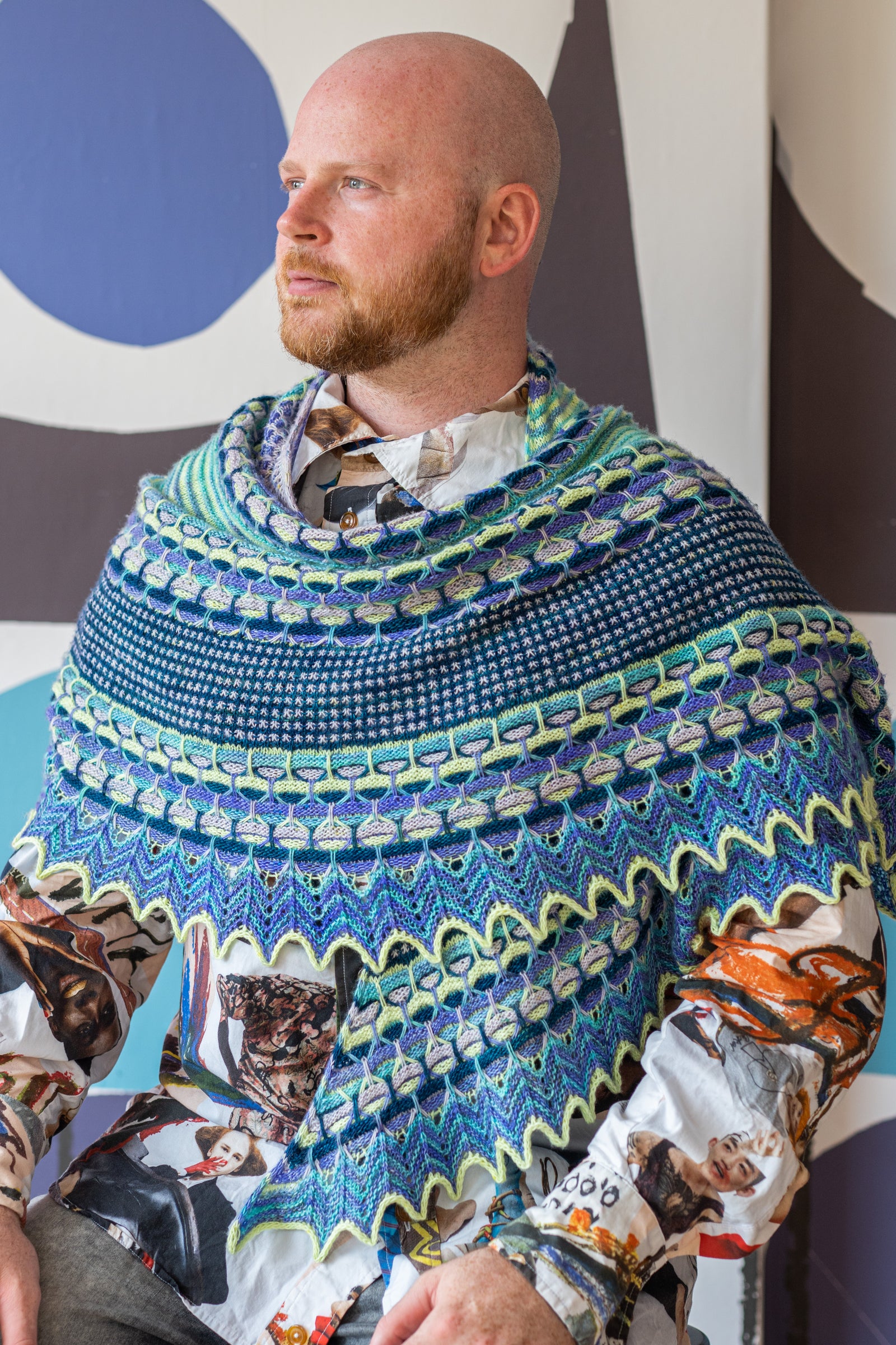 This image shows Stephen West wearing a beautifully knitted sample of the Aurora Cabin Shawl. The sample is knitted in blue, turquoise and purple shades. He stands on a colourful background. 