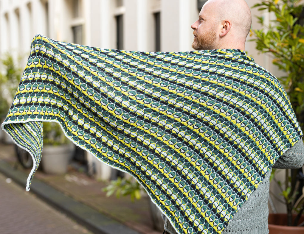 This image shows Stephen West wearing the  Aurora Cabin Triangle The shawl is knitted with green, yellow, and blue tones. The background shows a street in Amsterdam centre. 
