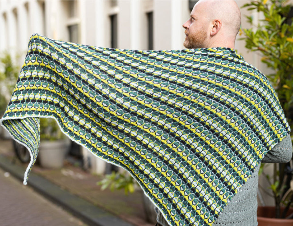 This image shows Stephen West wearing the  Aurora Cabin Triangle The shawl is knitted with green, yellow, and blue tones. The background shows a street in Amsterdam centre. 