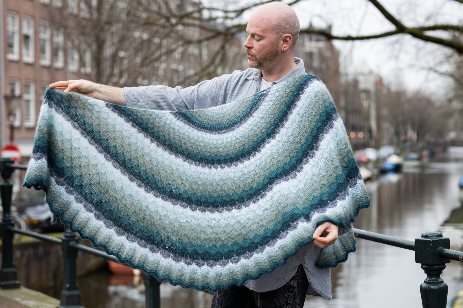 This image shows Stephen West holding a beautifully knitted shawl in cool, blue tones. The background shows a canal in Amsterdam centre. The shawl is named "The Awesomer Gossamer" and it was launched as part of the "Westknits Glow Along". 