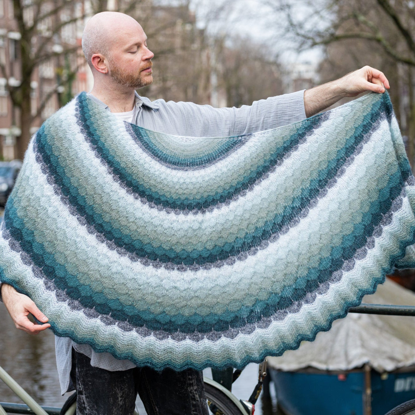 This image shows Stephen West holding a beautifully knitted shawl in cool, blue tones. The background shows a canal in Amsterdam centre. The shawl is named "The Awesomer Gossamer" and it was launched as part of the Glow Along.