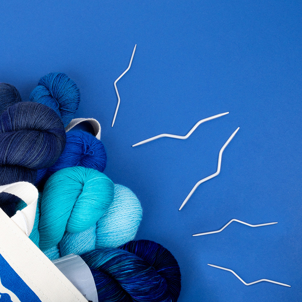 This image shows some cable needles on a blue background, laid next to some blue skeins of yarn. 