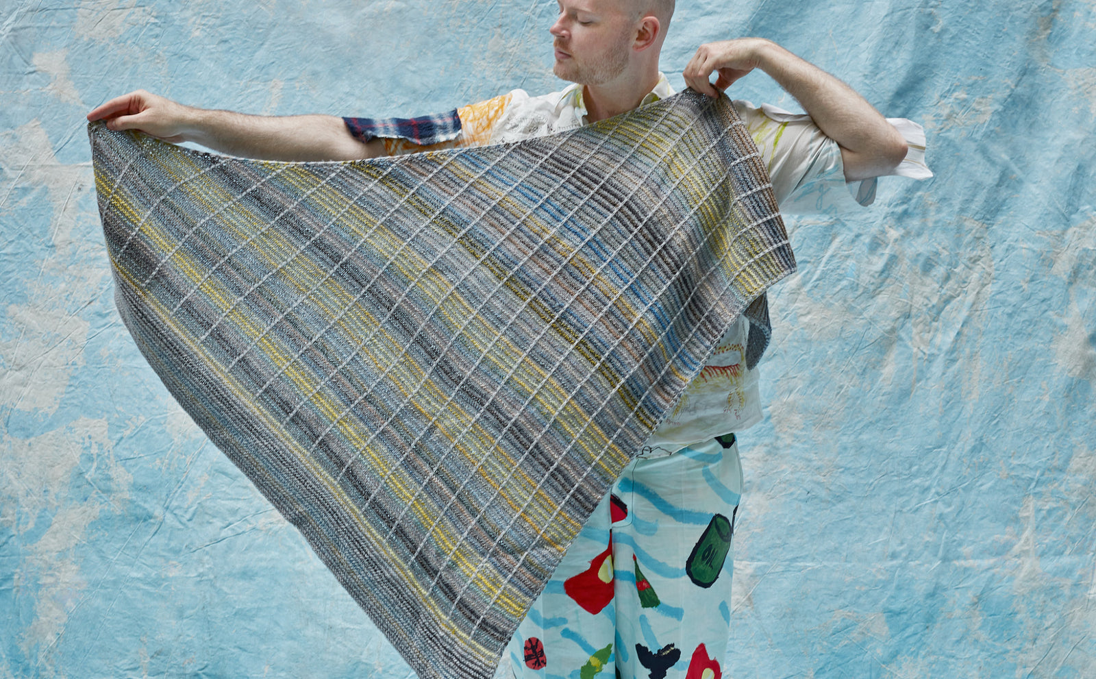 This image shows Stephen West holding a beautifully knitted sample of the Botanic Shawl by Westknits. The sample is knitted in yellow, grey, and blue pastel colours. It stands on a light blue background. 