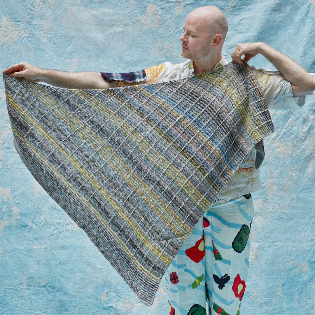 This image shows Stephen West holding a beautifully knitted sample of the Botanic Shawl by Westknits. The sample is knitted in yellow, grey, and blue pastel colours. It stands on a light blue background. 