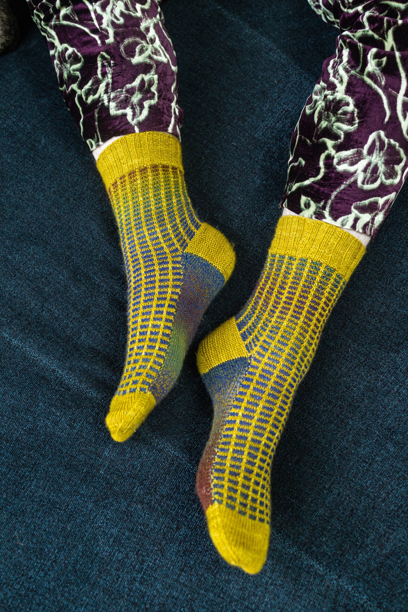 This image shows Stephen West wearing a yellow and blue sample of the Botanic Socks by Westknits. He stands on a denim blue background. 