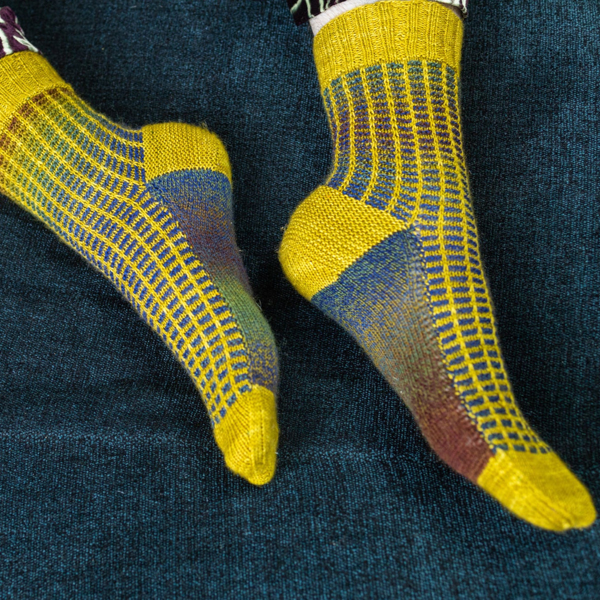 This image shows Stephen West wearing a yellow and blue sample of the Botanic Socks by Westknits. He stands on a denim blue background. 
